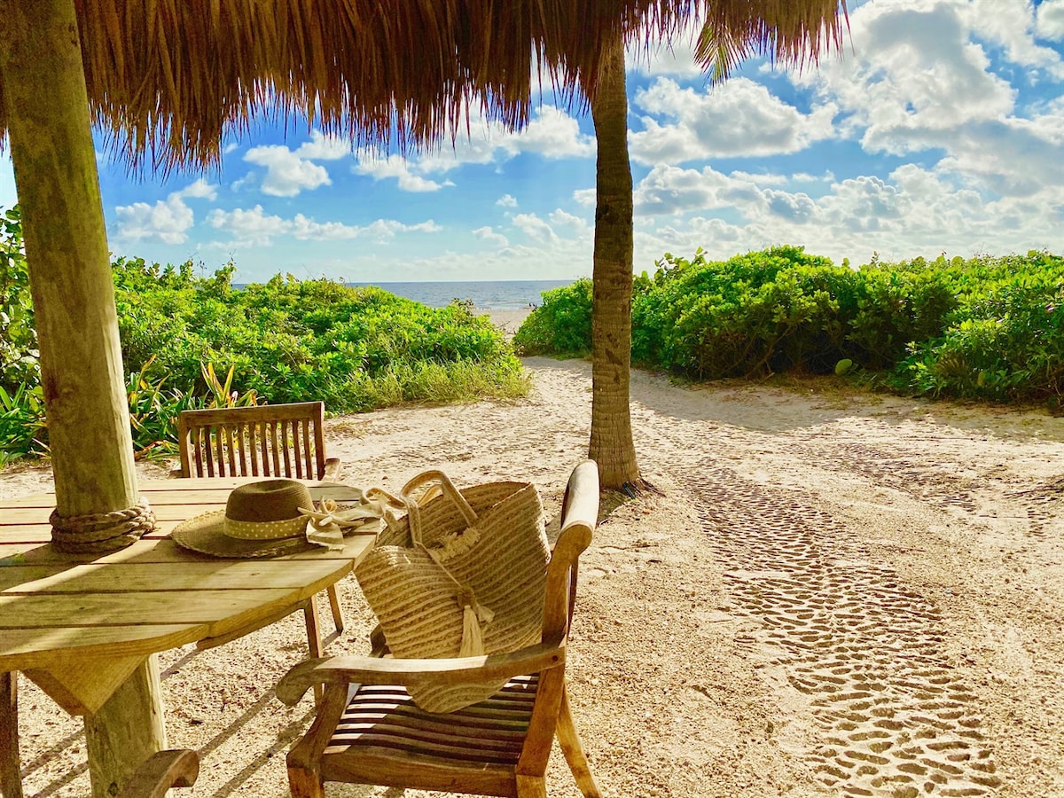 A shaded seating area is presented, featuring a wooden table and chairs. A straw bag and sunhat rest on the table, with a sandy path leading towards the beach visible in the background. Lush greenery and a partly cloudy sky enhance the serene setting.