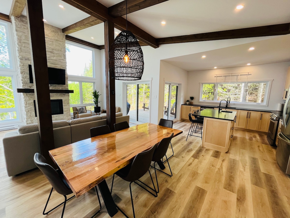 A spacious dining area features a wooden table surrounded by several chairs, with a stylish light fixture above. The open layout connects to a modern kitchen and a cozy seating area, illuminated by natural light pouring through large windows.