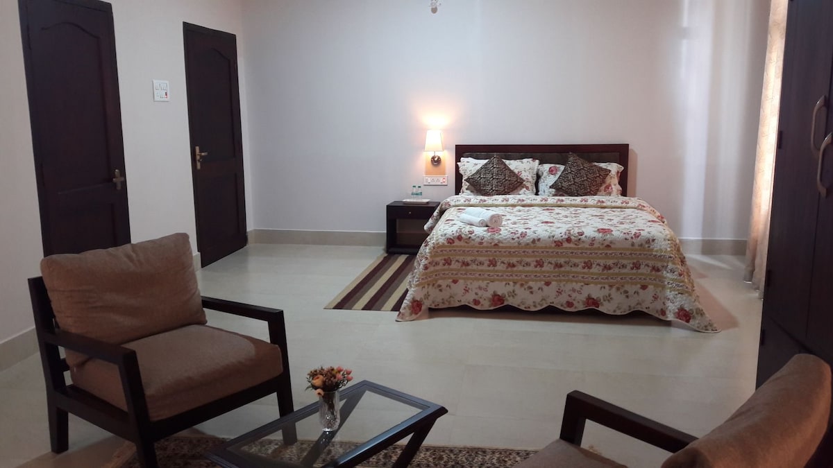 A cozy room features a spacious bed adorned with a floral-patterned coverlet and decorative pillows. Two comfortable chairs are positioned nearby, complemented by a small glass table. Soft lighting from a wall sconce creates a relaxed ambiance, while natural light filters through a window with sheer curtains.