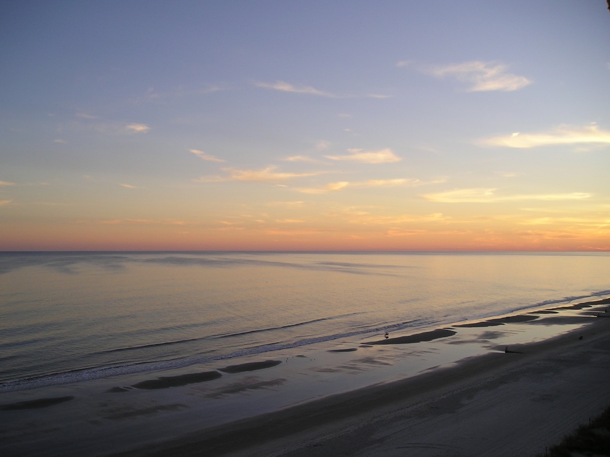 A serene ocean view captures the gentle waves lapping against the shore, under a sky painted with soft hues of orange and purple as the sun sets. The calm water reflects the colorful skyline, creating a tranquil atmosphere.