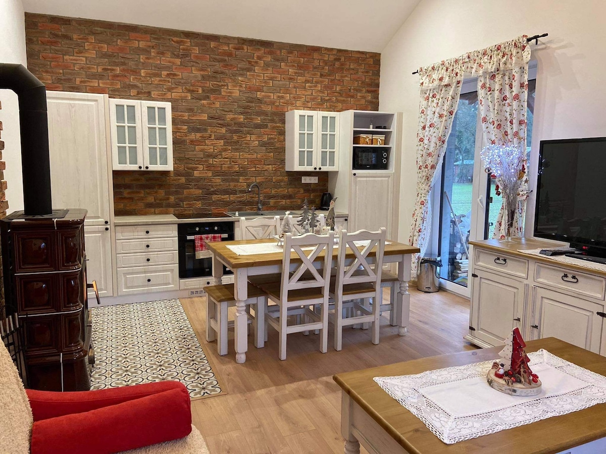 A well-equipped kitchen and dining area are shown, featuring a central wooden table surrounded by white chairs. A brick wall provides texture, while light-colored cabinets and appliances create a bright atmosphere. Decorative curtains frame the windows, and a cozy seating area is visible.