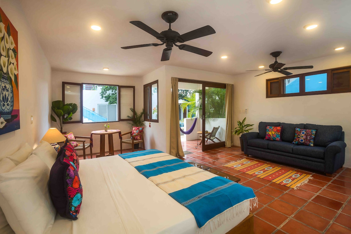 A spacious guest room is furnished with a large bed adorned with colorful pillows. A comfortable sofa is positioned nearby, and a small dining table is visible. Large windows provide natural light and views of the outdoor area, enhancing the inviting ambiance.