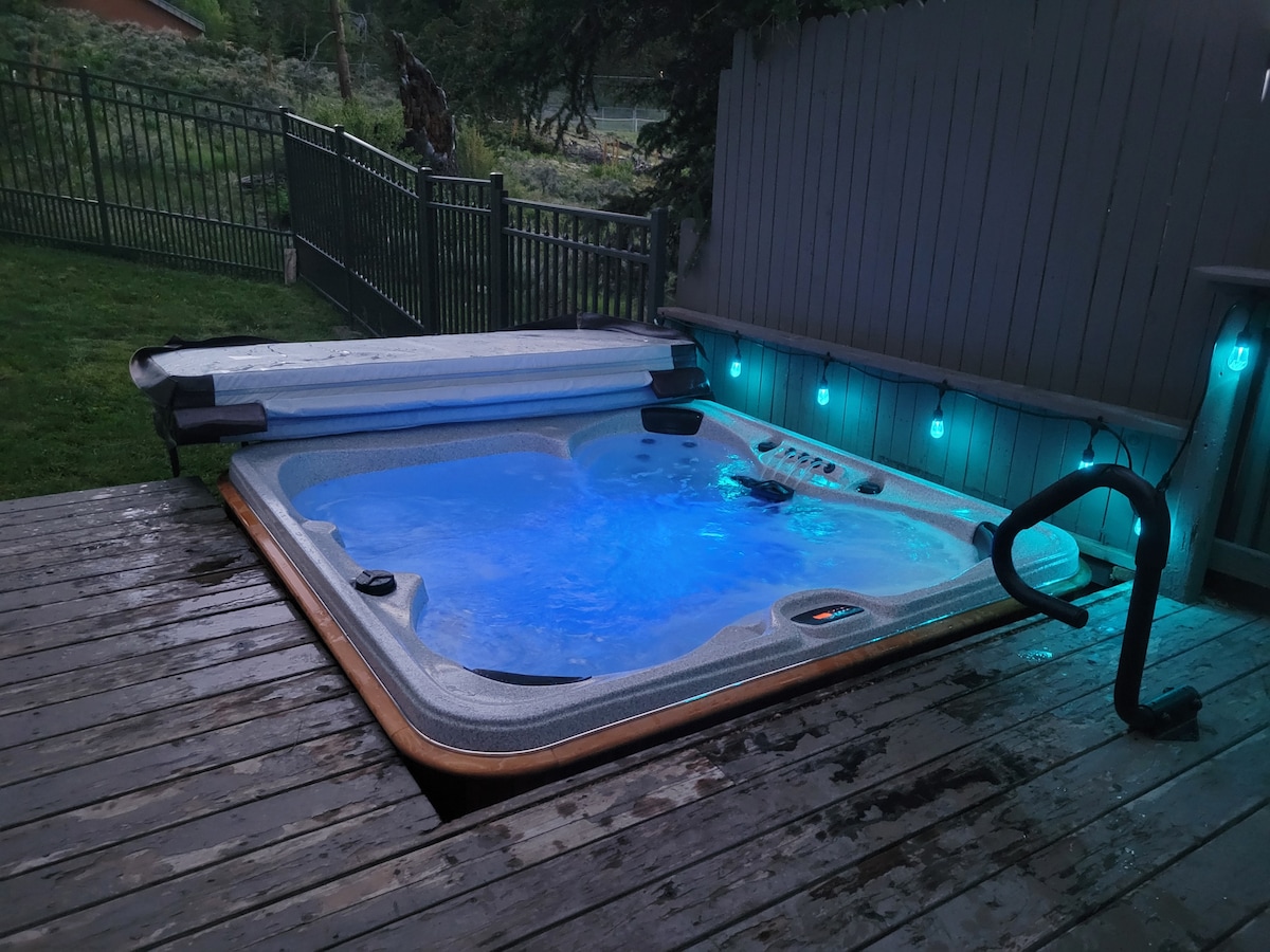 A hot tub is positioned on a wooden deck, surrounded by a grassy area and a fenced yard. Soft blue lighting enhances the ambiance, with water visible inside the tub. A safety railing is included for ease of access.