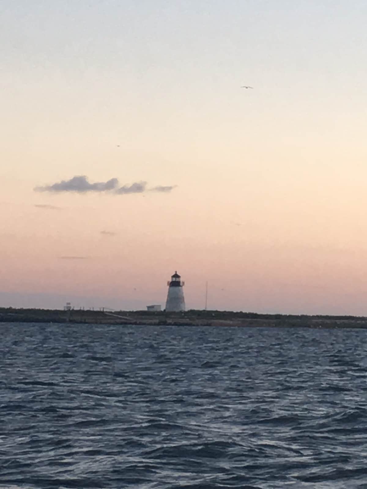 A distant lighthouse is seen against a soft pastel sky during twilight. The ocean waves gently ripple in the foreground, reflecting the fading light of the day. The scene conveys a peaceful coastal atmosphere.