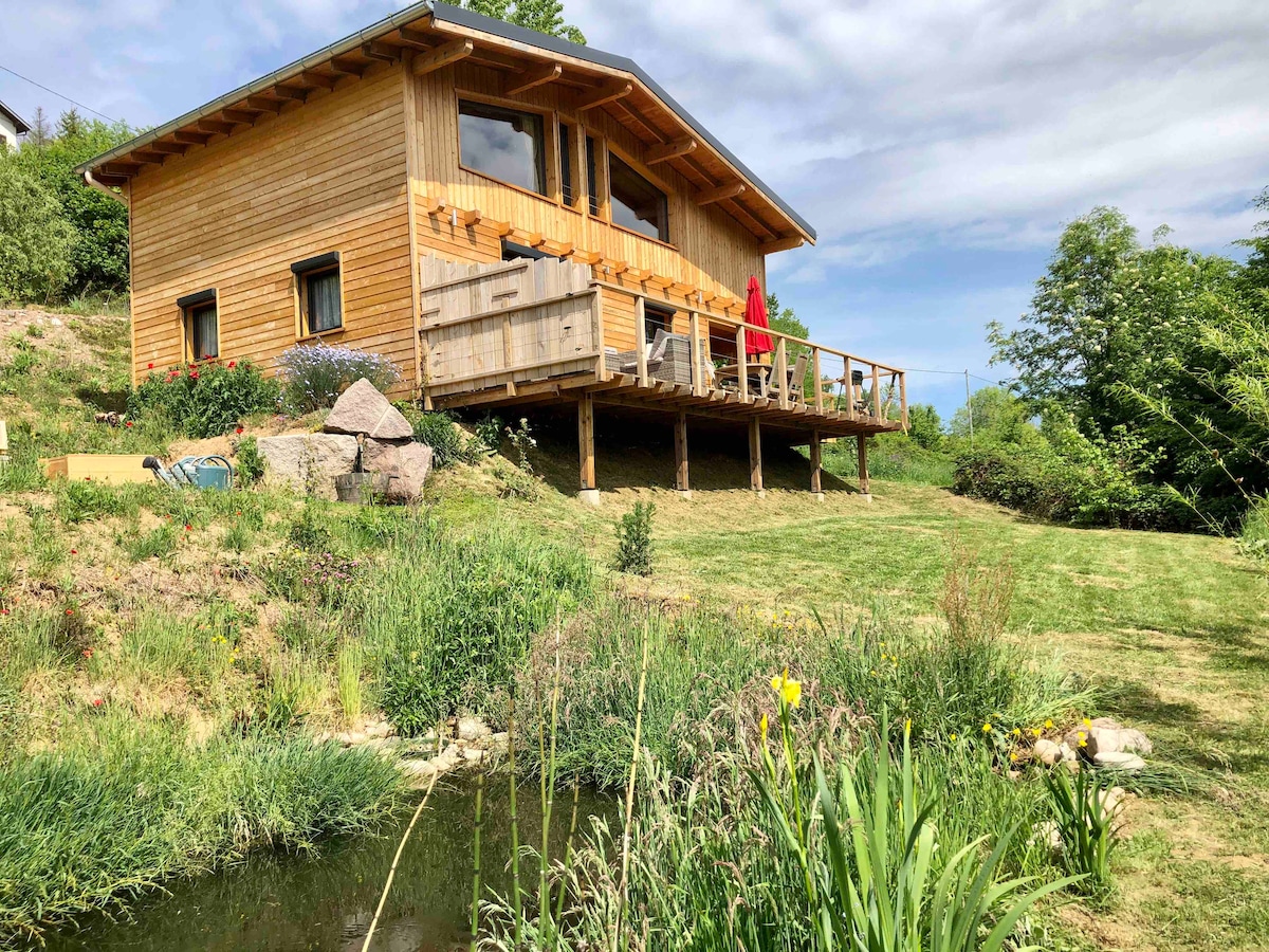 The bioclimatic wooden chalet is positioned on a sloped terrain, featuring large windows and a spacious deck adorned with outdoor seating. Surrounding greenery includes well-maintained grass and various plants, complementing a serene pond with reeds and flowers along the water’s edge.
