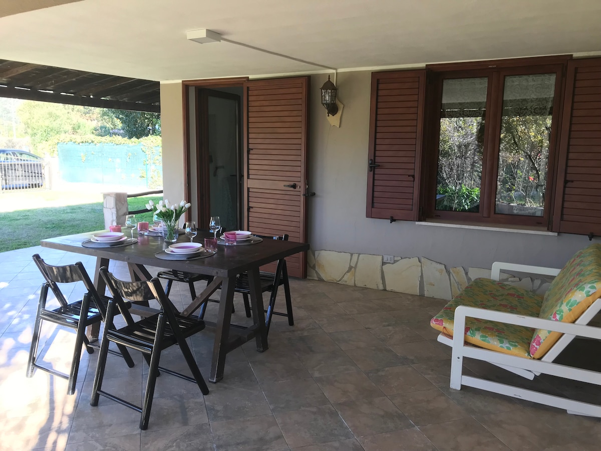 An outdoor dining area is present under a covered porch, featuring a wooden table set for meals with plates and glasses. Relaxing seating is provided by a cushioned sofa. Large windows allow natural light, and the surrounding garden offers a serene view.