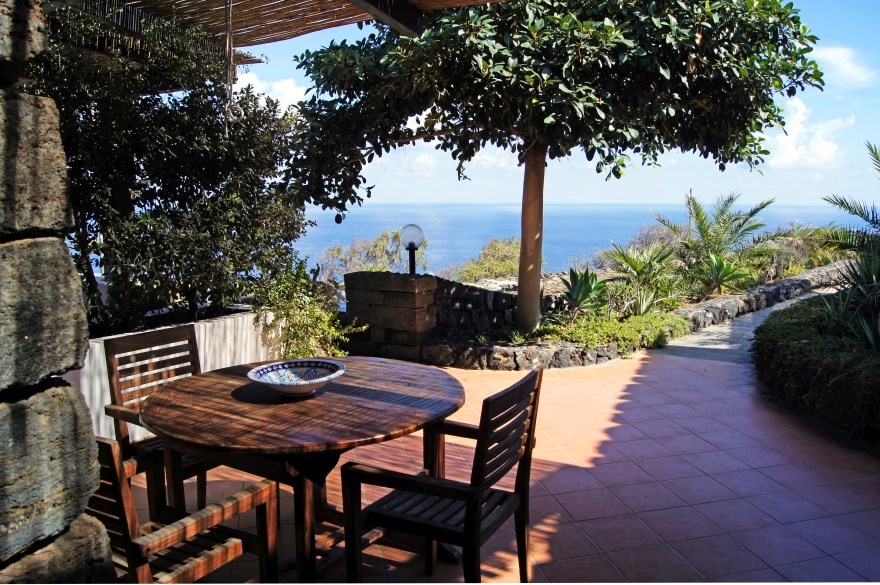 An outdoor terrace features a round wooden table surrounded by four chairs, offering a space for al fresco dining. Lush greenery and gently sloping paths lead toward the horizon, where a glimpse of the sea can be seen under a clear sky.