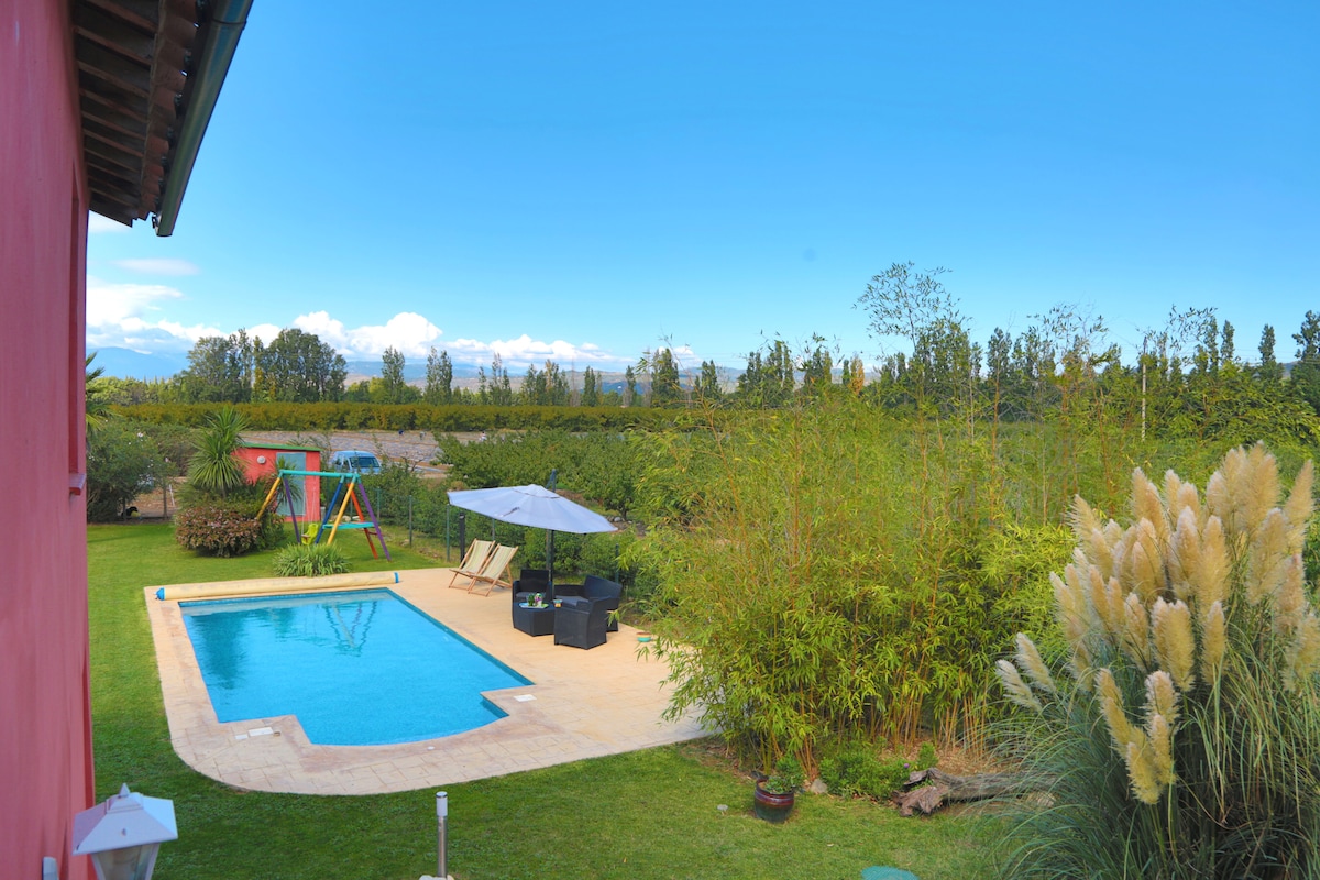 A serene outdoor space features a sparkling pool surrounded by lush greenery. Comfortable seating is arranged nearby, complemented by an umbrella for shade. A playset stands in the background, all set against a clear blue sky and distant mountains.
