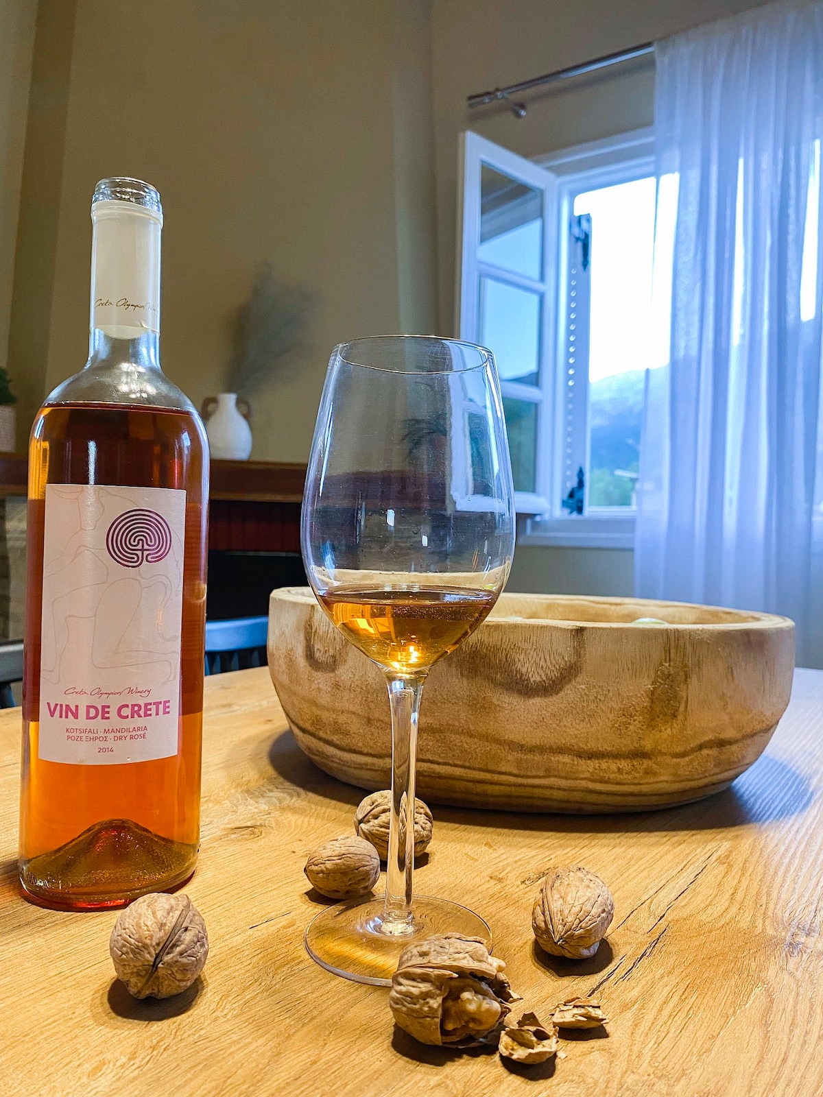 A glass of Cretan wine sits on a wooden table, accompanied by walnuts scattered nearby. A bottle labeled 'Vin de Crète' is prominently displayed. Natural light streams through an open window, highlighting the warm tones of the wooden bowl in the background.
