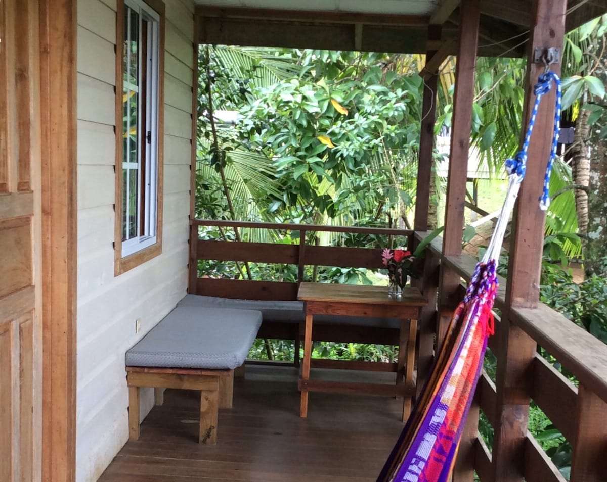 A shaded balcony area features a cozy bench with a light cushion and a small wooden table. A colorful hammock is draped nearby, surrounded by lush green foliage. A small vase with flowers adds a touch of warmth to the space.
