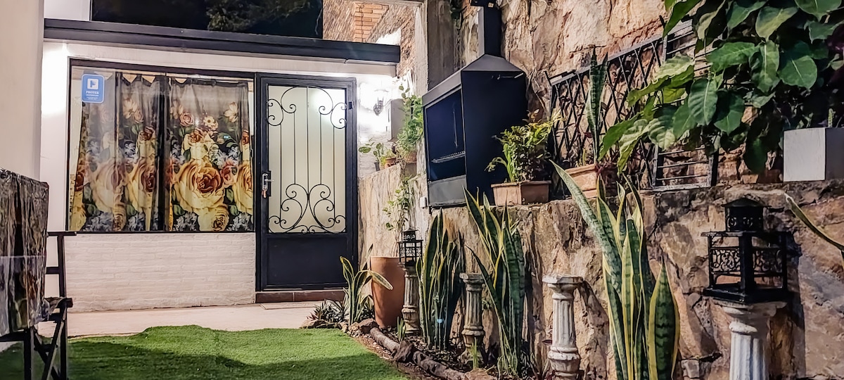 An entryway is depicted, featuring a decorative door adorned with floral patterns. Surrounding greenery and decorative plants create a welcoming atmosphere. Outdoor seating is arranged to the side, complemented by lanterns and a small table. The stone wall adds a rustic charm to the space.