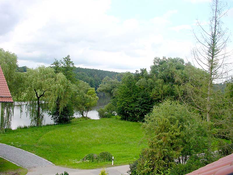 A serene landscape view features a lush green area bordered by tall trees, overlooking a calm river. The sky is partly cloudy, and a winding pathway can be seen leading towards the water. Natural beauty surrounds the space, inviting leisurely outdoor activities.