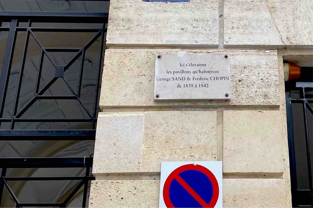 A stone wall displays a plaque honoring George Sand and Frédéric Chopin, indicating their residence from 1839 to 1842. Below, a no parking sign is visible, contributing to the urban context. The architectural details of the building are framed by distinct ironwork.