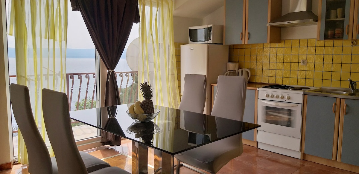 A modern kitchen area features a sleek glass dining table surrounded by four light-colored chairs. Yellow drapes frame the window, allowing natural light to illuminate the space. The kitchen is equipped with essential appliances and offers a view of the sea through the balcony doors.