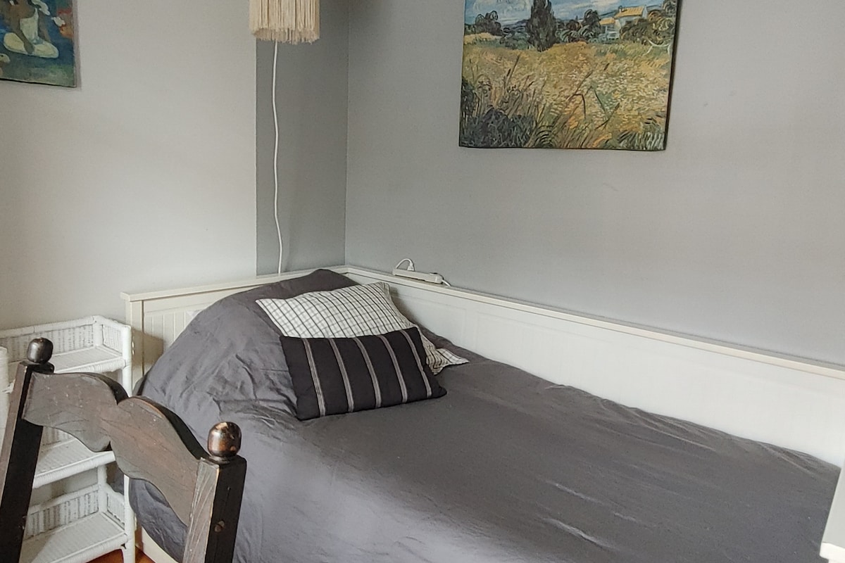 A simple bedroom features a spacious bed dressed in dark bedding, complemented by decorative cushions. A small bedside table is positioned nearby, along with a chair visible in the foreground. The walls are painted in a soft grey tone, adorned with a landscape painting that adds a touch of color.
