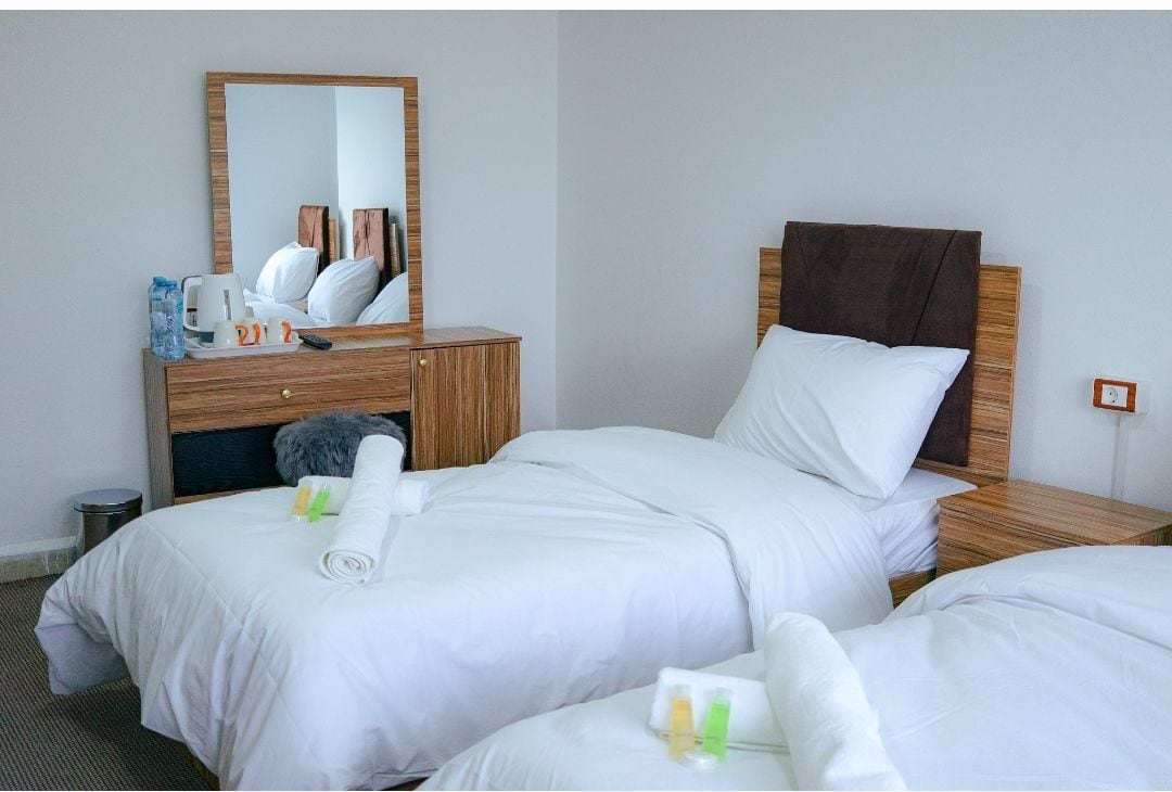 A room features two twin beds dressed in white linens, complemented by wooden headboards. A mirror above a matching chest of drawers reflects the bright space. Toiletries and towels are neatly arranged on each bed, creating a tidy and welcoming environment.