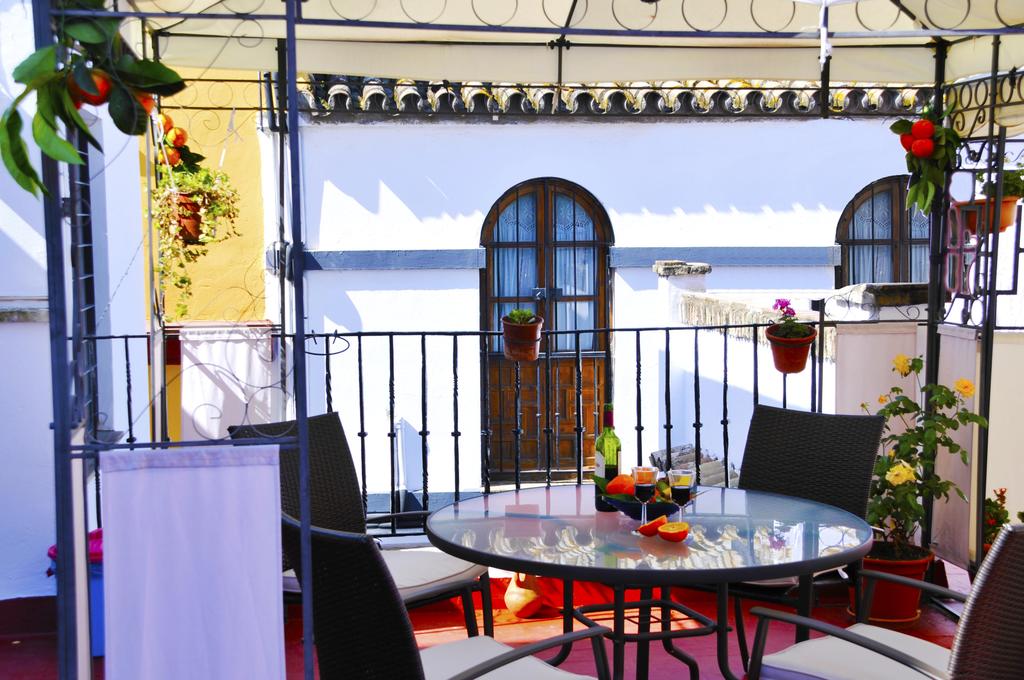A terrace area features a round glass table surrounded by four dark wicker chairs. Potted plants are attached to the railing and walls, while a bottle and glasses are placed on the table. A large window with a dark wooden frame is visible in the background.