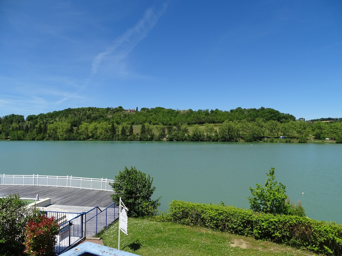 A serene view of the lake is displayed, bordered by lush greenery and trees. The calm water reflects the clear blue sky, with a wooden dock visible along the shoreline, offering a tranquil setting for relaxation by the water.