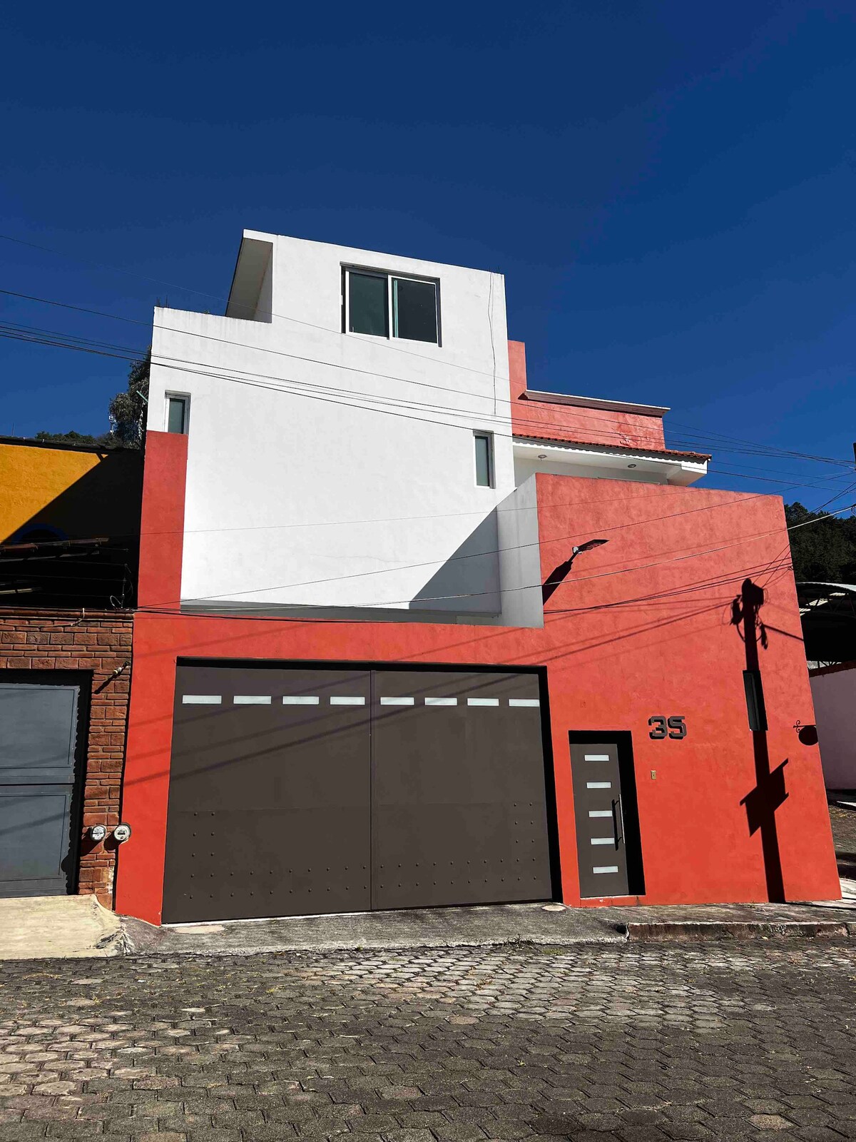 The exterior of a modern three-story building is presented, featuring a combination of orange and white walls. A large garage door is visible at ground level, accompanied by a contemporary front door. A window illuminates the upper level, reflecting the clear blue sky.