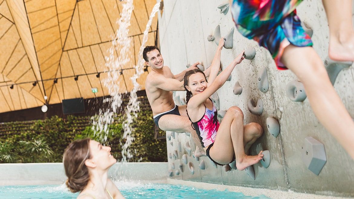 A climbing wall integrated into a pool area features individuals engaging in climbing activities. Water cascades from above, creating an inviting atmosphere. The setting includes a spacious pool with light blue water, surrounded by a warm and modern architectural design.