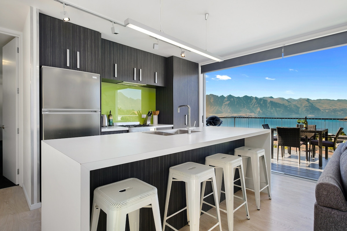 A modern kitchen area is seen, featuring sleek black cabinetry and a spacious white countertop. Stainless steel appliances, including a refrigerator, are integrated seamlessly. Large glass doors reveal a stunning view of mountains and a lake, enhancing the connection between the indoor and outdoor spaces.