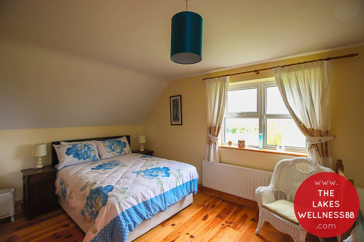 A bright bedroom features a spacious bed adorned with floral bedding. Two bedside lamps sit atop dark wooden nightstands. Natural light enters through a large window, framed by light curtains. The warm wooden floor complements the soft cream walls, creating a welcoming environment.