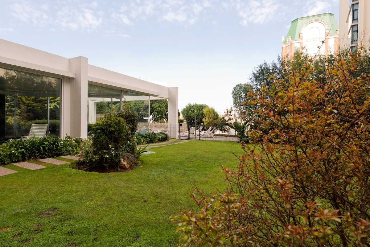A landscaped garden area features well-maintained grass and various shrubs, creating a serene outdoor space. The property building is visible in the background, showcasing large glass windows that invite natural light into the interior. Pathways lead through the greenery, enhancing accessibility.