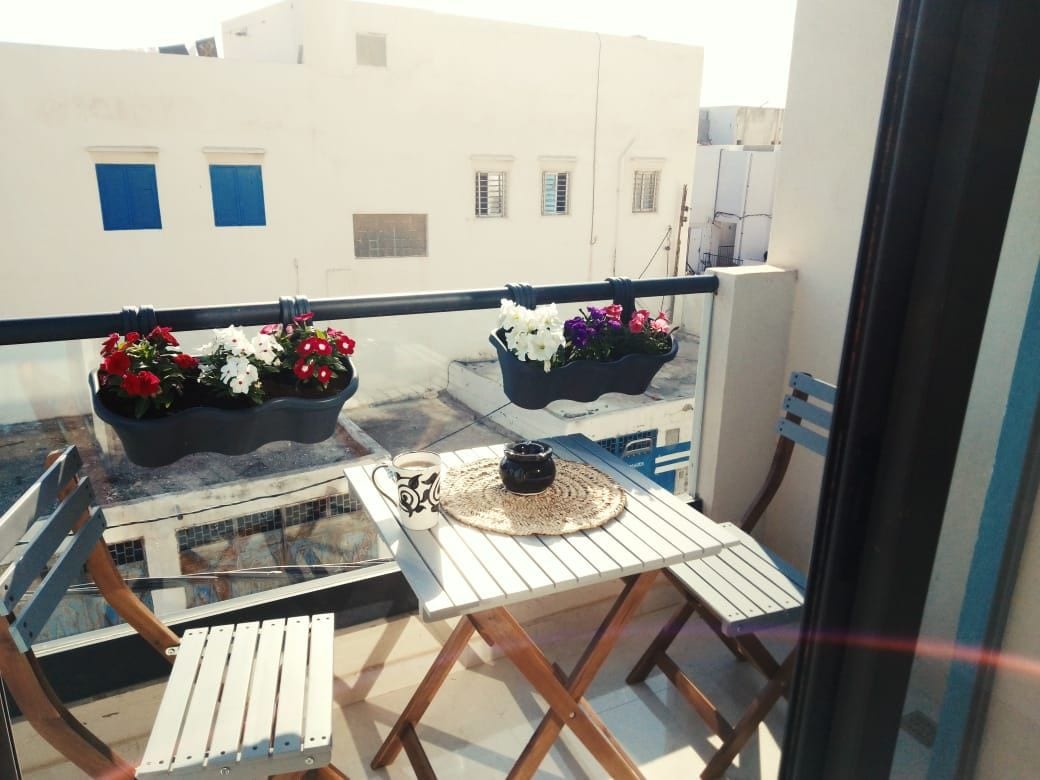 A small balcony is furnished with two wooden chairs and a table, adorned with a round placemat and a decorative bowl. Flower boxes hang from the railing, filled with vibrant red and white blooms, creating a welcoming outdoor space.