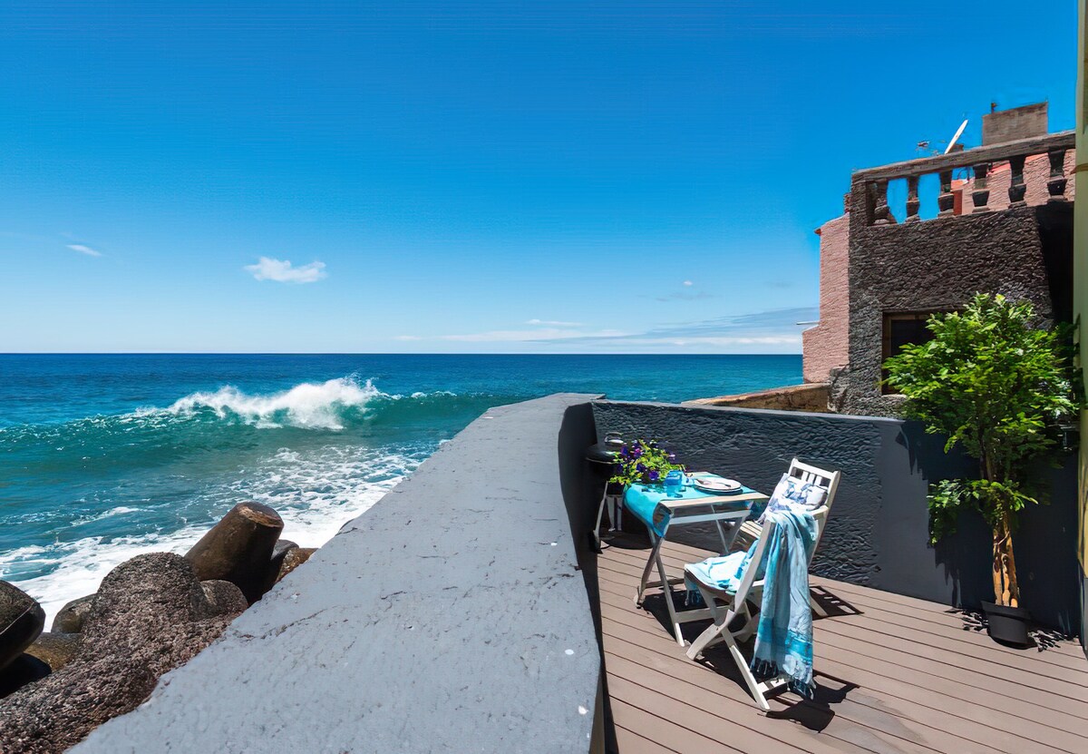 A scenic terrace is seen overlooking the ocean, with gentle waves visibly crashing against the nearby rocks. A small table is set with a blue tablecloth, flanked by two white chairs. Lush greenery is positioned beside the seating area, adding a touch of nature to the coastal view.