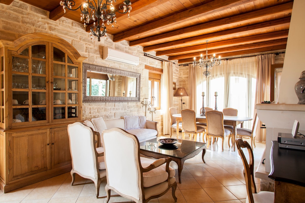 The lounge area features exposed wooden beams and stone walls, creating a warm ambiance. Elegant seating is arranged around a glass-top table, complemented by a stylish wooden cabinet and soft drapes that frame the windows. Decorative light fixtures enhance the inviting atmosphere.