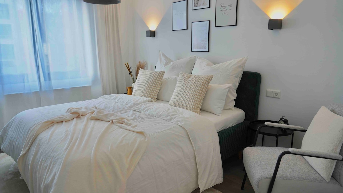 A cozy bedroom features a spacious bed adorned with soft white linens and decorative pillows. Natural light enters through a large window, which is complemented by minimalistic wall art and stylish sconces. A comfortable chair and small side table add to the inviting ambiance.