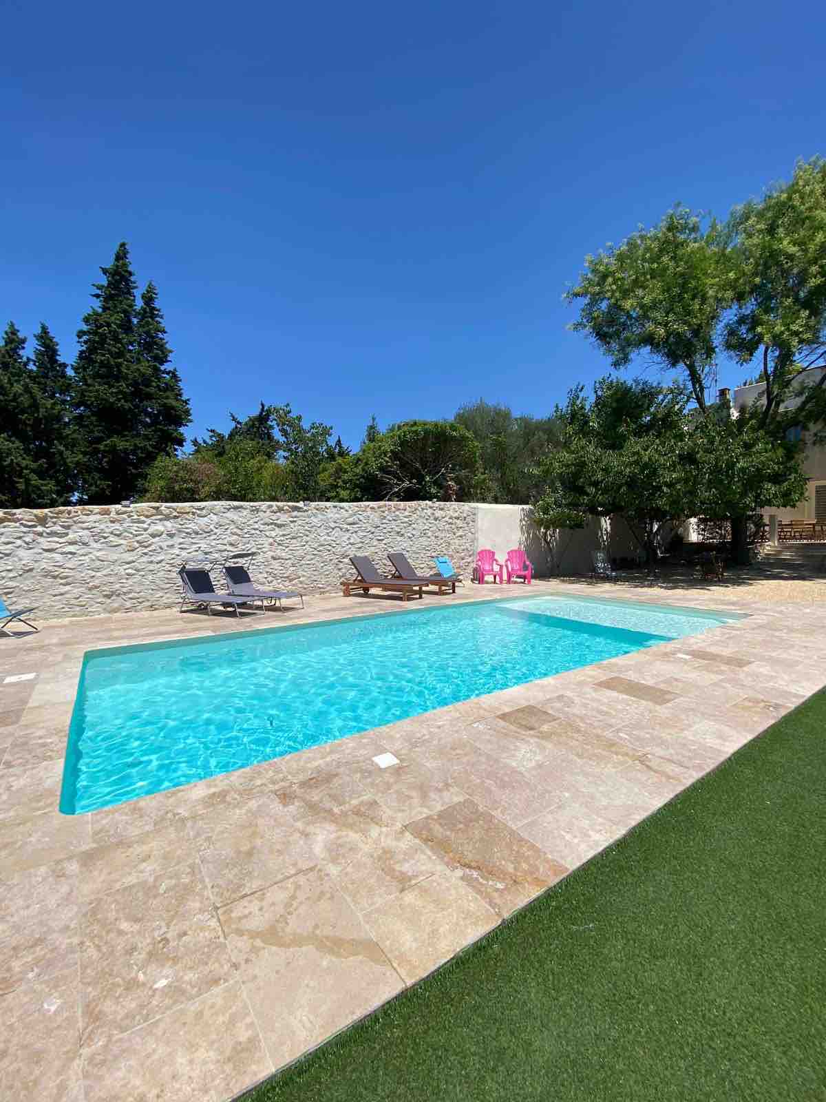 A refreshing swimming pool is set against a stone wall, surrounded by a well-maintained outdoor area. Sun loungers in neutral tones are positioned alongside the pool, and vibrant lounge chairs are placed nearby, providing ample seating options. Lush greenery is visible in the background.