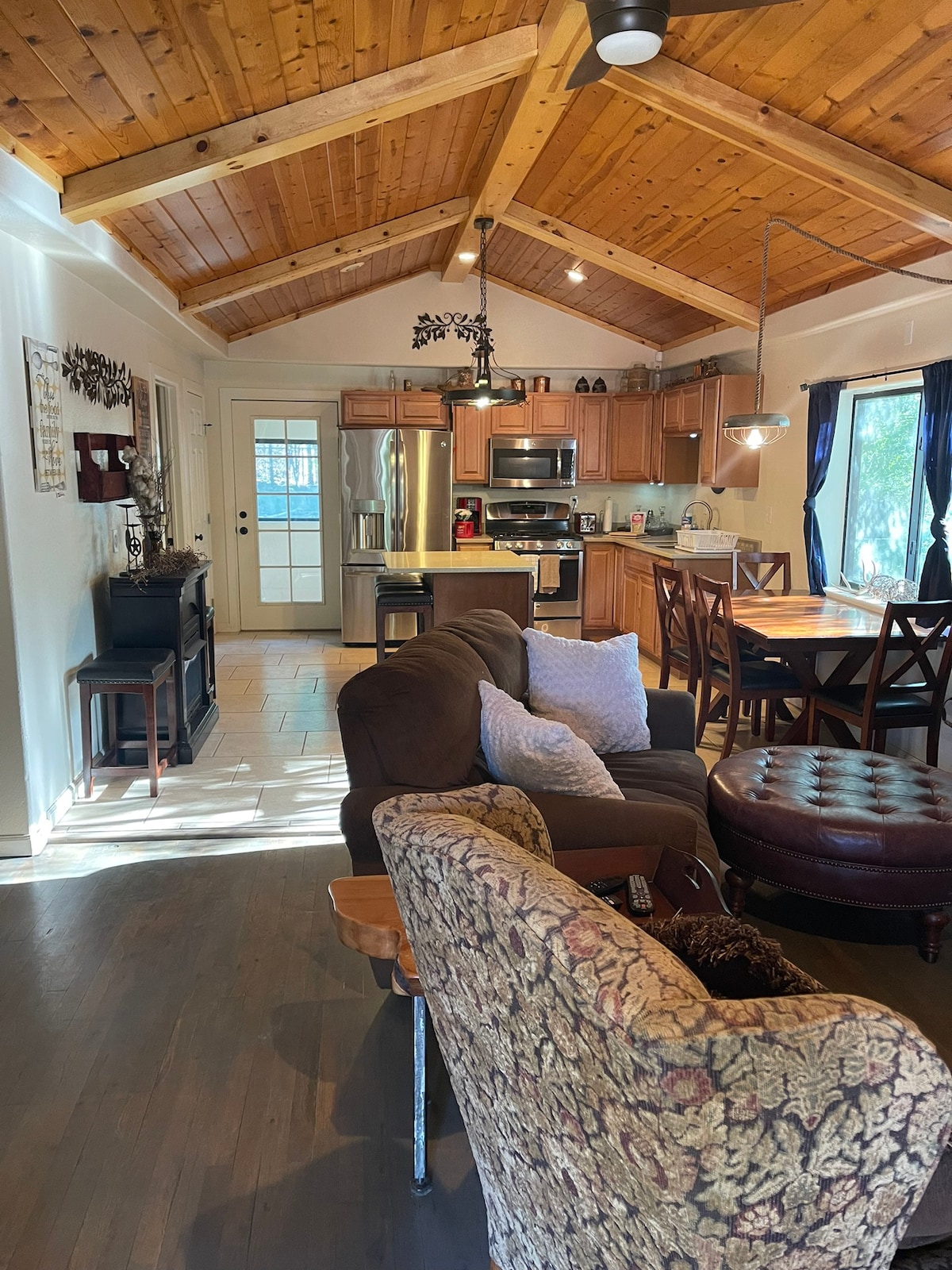 An inviting interior showcases a spacious living area with a mix of comfortable seating options. A wooden ceiling with exposed beams offers a warm feel, while the kitchen and dining area feature modern appliances and natural light filtering through large windows.
