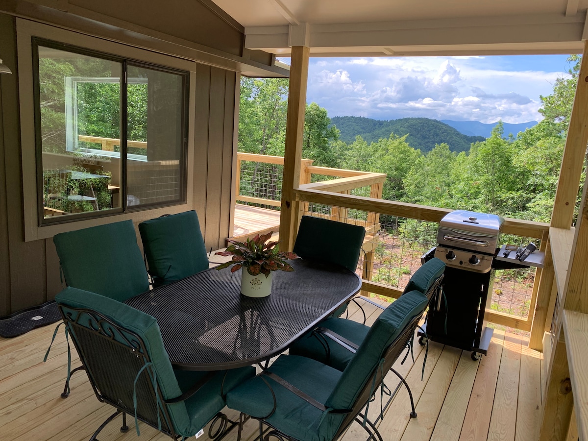 An outdoor dining area is situated on a spacious deck, featuring a table with six cushioned chairs. A gas grill stands nearby, with lush green trees and distant mountains visible in the background under a partly cloudy sky.