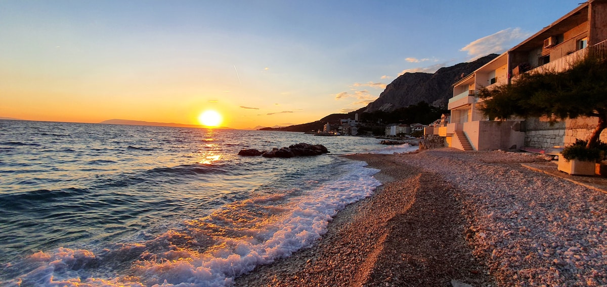 The shoreline of a pebble beach is displayed, with gentle waves lapping against the rocks. A vibrant sunset paints the sky in warm hues, casting a soft glow over the water. In the background, the outline of a coastal village and hills is subtly visible.