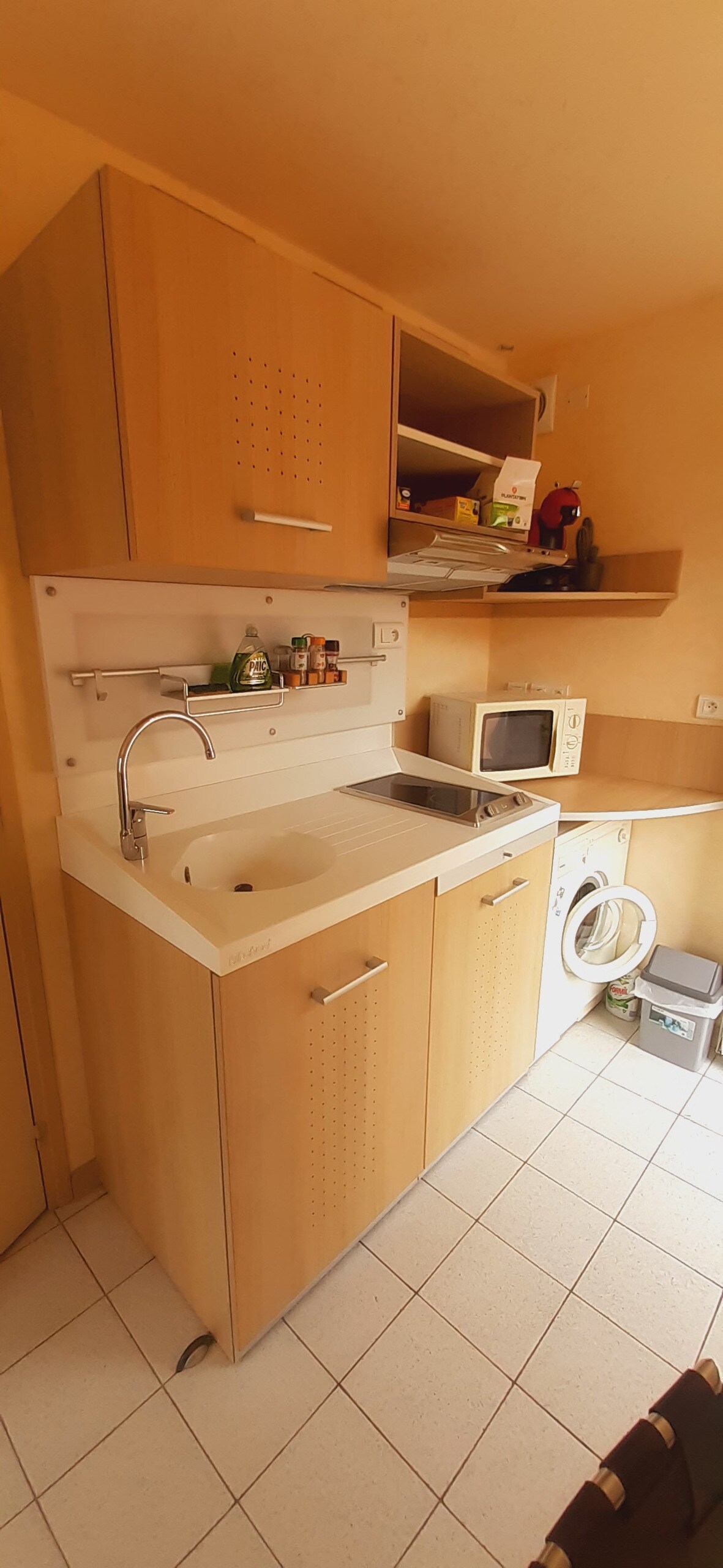 A compact kitchenette features light wood cabinetry with a sink and a small countertop. A microwave and a washing machine are positioned below upper shelves that store various items. Bright tiles cover the floor, enhancing the functionality of the space.