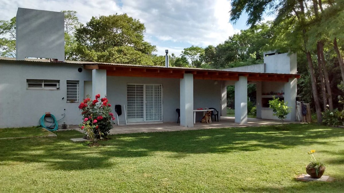 A single-story house surrounded by greenery features a spacious covered patio with wooden beams. Large glass sliding doors provide access to the interior. Colorful flowers are planted in the foreground, adding a natural touch to the outdoor space, which is well-maintained and inviting.