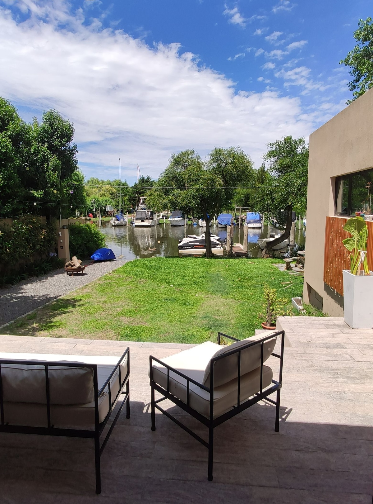 A serene outdoor area features comfortable seating, providing a view of the waterway and boats. Lush greenery surrounds the space, and a pathway leads to the waterfront, enhancing the connection between nature and the living area.