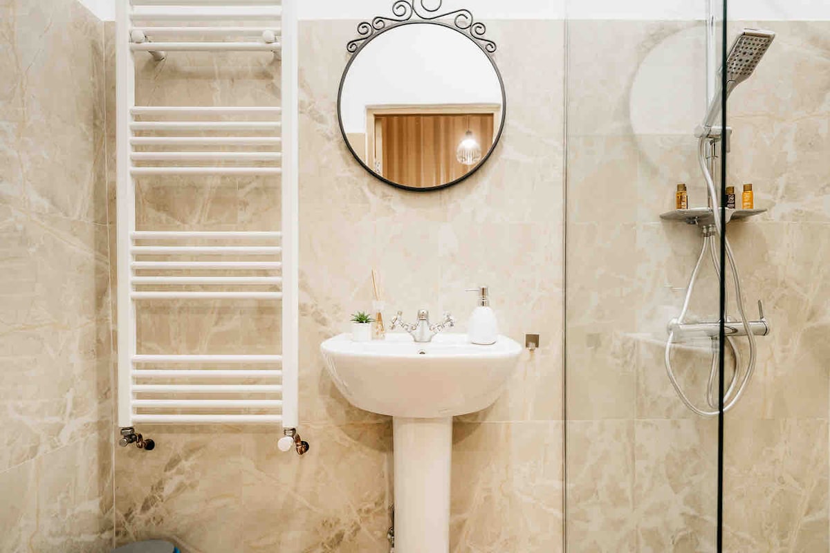 The bathroom features marble-patterned walls, a modern pedestal sink with a round mirror above. A shower area is enclosed by glass, and a sleek showerhead is mounted on the wall. A heated towel rack is positioned to the side, adding functionality.