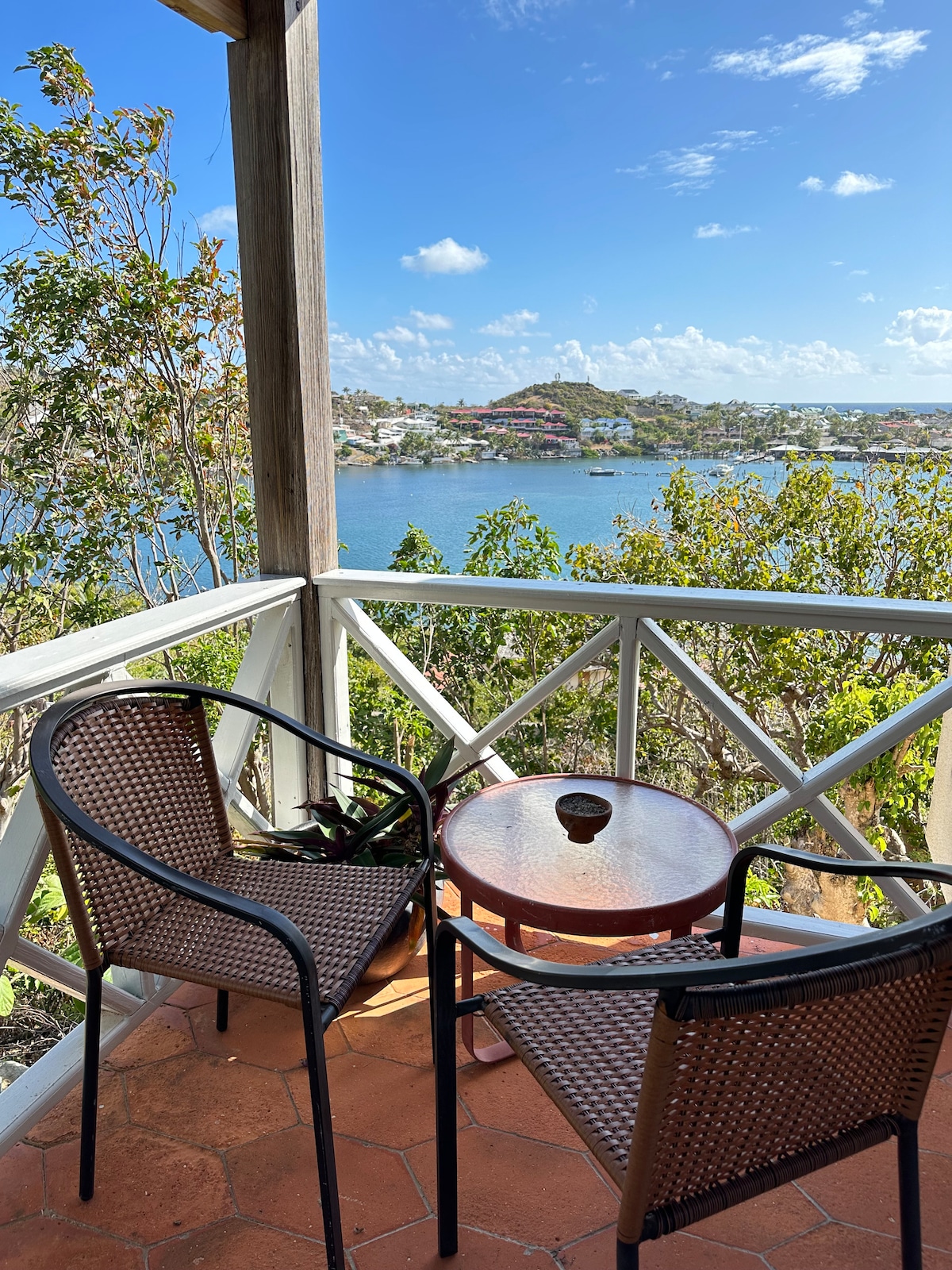 A charming balcony is furnished with two wicker chairs and a small round table, offering a serene setting. Lush green foliage surrounds the area, while a picturesque view of the pond and distant hillside is visible under a clear blue sky.