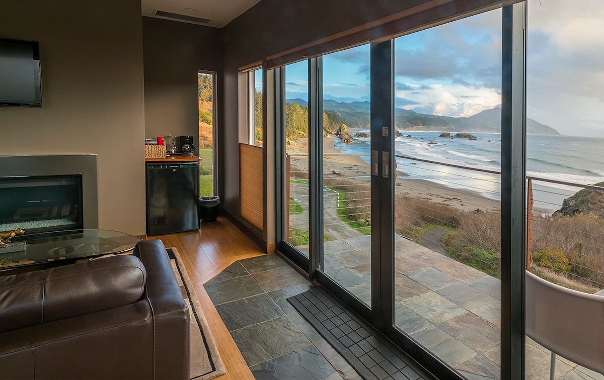 A contemporary living space features large glass doors offering unobstructed views of the ocean and beach. A plush leather sectional is positioned near a sleek gas fireplace. Natural light fills the area, enhancing the connection between indoors and the beautiful coastal scenery.