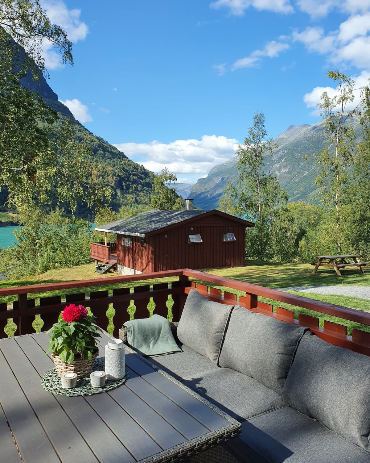 A welcoming outdoor seating area features a grey upholstered sofa and a wooden table, complemented by a potted flowering plant. In the background, a cozy red cabin is positioned amidst lush greenery and mountainous terrain, with a glimpse of the turquoise water visible beyond.