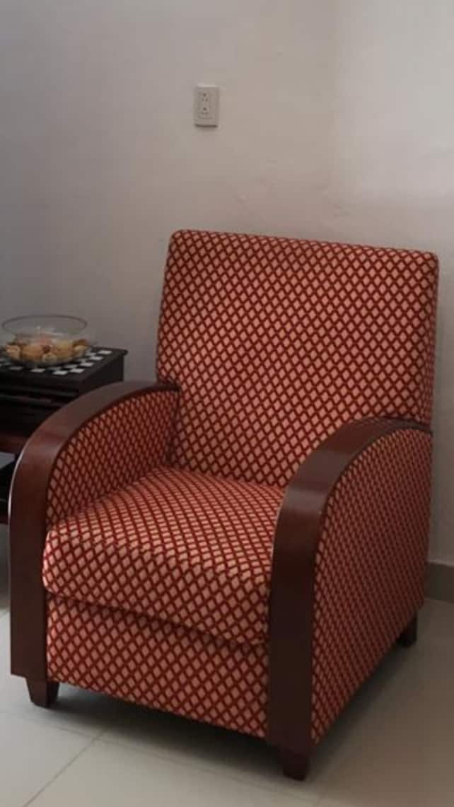 A patterned armchair with a wooden frame is positioned in a corner. The fabric features a diamond design in warm shades of orange and cream. Beside it, a small table holds a decorative bowl filled with assorted fruits.