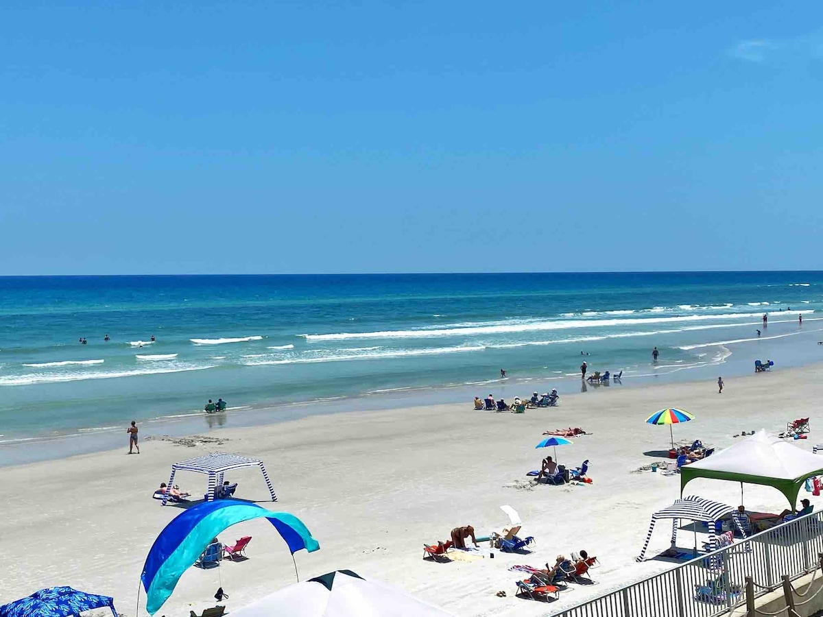 A wide view of a sandy beach is offered, showcasing families and individuals enjoying the sun and surf. Colorful umbrellas dot the shore, and gentle waves lap at the shore under a clear blue sky. The scene captures a serene beach atmosphere.