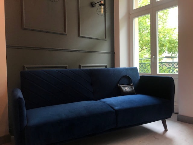 A deep blue sofa with a textured fabric is positioned against a dark accent wall. Natural light enters through a window, framed by a black iron balcony railing, creating a soft contrast with the surrounding neutral tones.