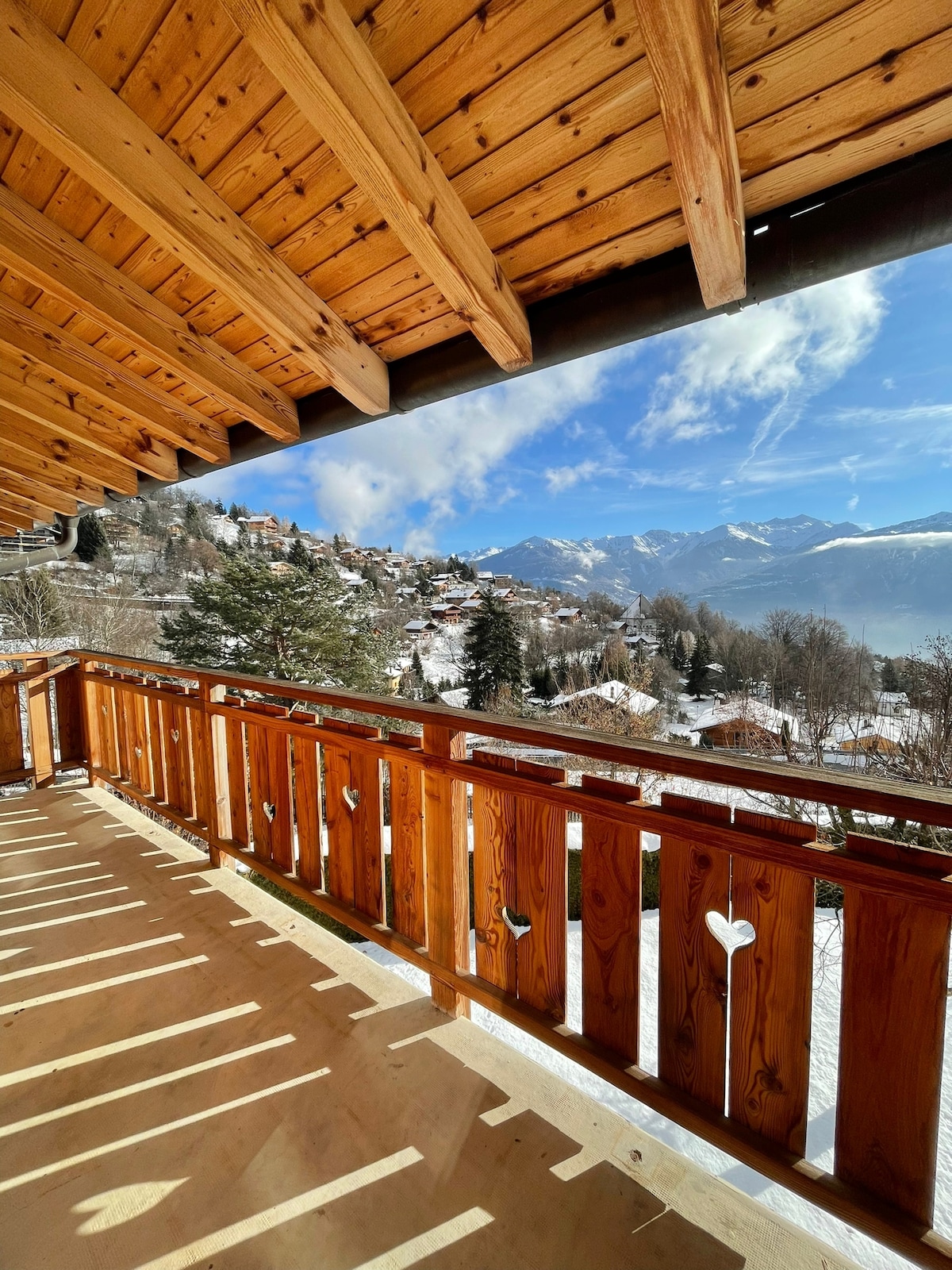 A wooden balcony adorned with a decorative railing overlooks a scenic view of snow-covered mountains and a hillside village. The warm wooden beams of the ceiling create a cozy ambiance, while the sunlight casts gentle shadows on the balcony floor.