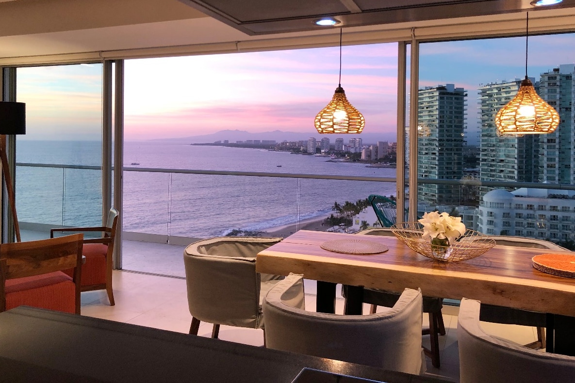 A dining area with a wooden table is illuminated by pendant lights, offering a view of the ocean and nearby buildings through expansive glass walls. Soft seating surrounds the table, while the vibrant colors of the sunset reflect off the water.