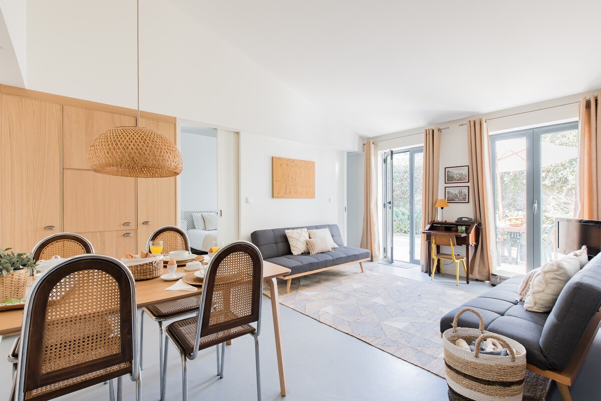 A spacious living area features a light-filled design with large glass doors overlooking an outdoor terrace. A dining table with woven chairs sits adjacent to a cozy seating area. Neutral tones and contemporary decor create a harmonious and relaxing environment.