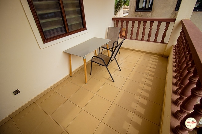 A small terrace features a light-colored table and two black chairs, creating an inviting space for relaxation. The area is surrounded by a wooden railing and tiled flooring, which enhances the simplicity of the outdoor setting.