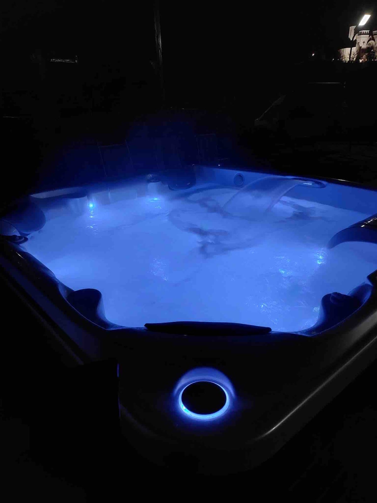 A hot tub is showcased, with soft blue lighting illuminating the water. Bubbles can be seen swirling, adding to the serene atmosphere. The surrounding area is dimly lit, enhancing the tranquil setting for relaxation.